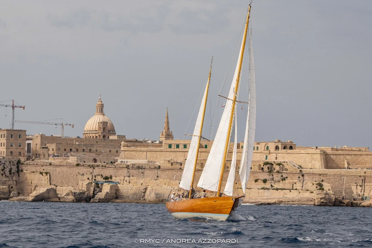 A varnished wooden schooner under white sails beneath the fortifications of Valletta, a domed church and spire on the skyline under a grey sky (2025).