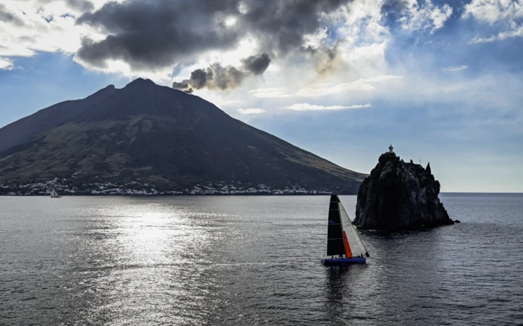 A racing yacht with dark sails passes the rock of Strombolicchio, the cone of Stromboli rising behind under broken cloud (2025).