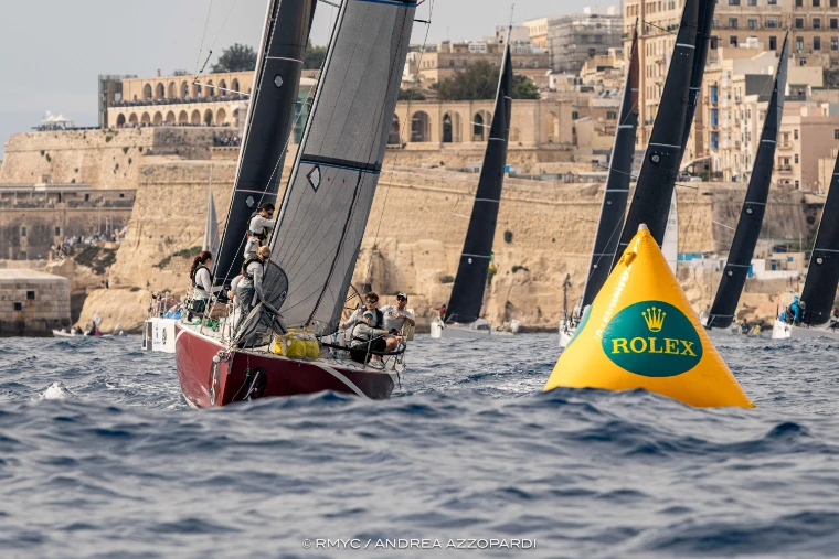 A red-hulled yacht with its crew hiked out passes a yellow Rolex buoy, other yachts under dark sails beyond, beneath the bastions and arcades of Valletta (2025).