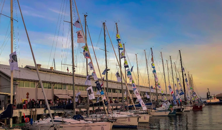 Race yachts dressed with flags lie moored in a row along a Hobart wharf at sunset, reflected in the still water (2018).