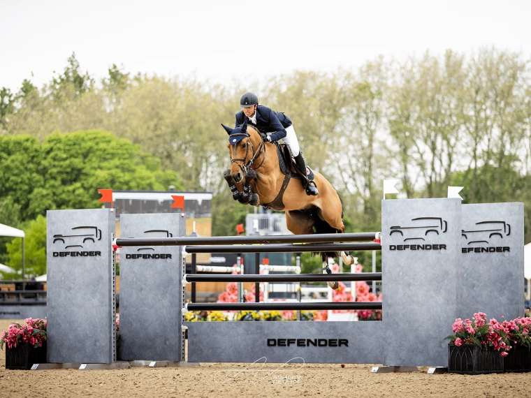A chestnut horse and rider clear a grey oxer in the international show jumping arena, pink flowers at its foot. The Royal Windsor Horse Show in the Home Park, Windsor, in the season calendar of The Mansa Court