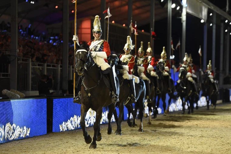 Mounted troopers of the Household Cavalry in plumed helmets ride past the stands in the floodlit evening arena, in 2024. The Royal Windsor Horse Show in the Home Park, Windsor, in the season calendar of The Mansa Court