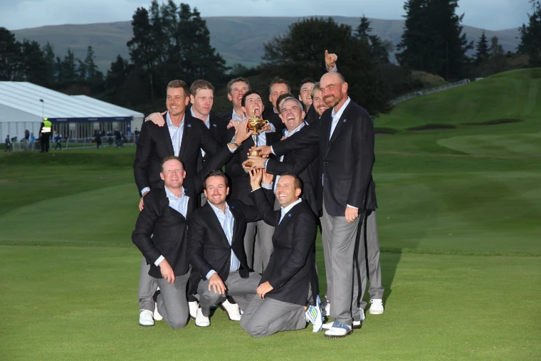 A team of golfers in dark blazers and grey trousers crowd together on the course, laughing and holding up the gold Ryder Cup at dusk (2014).