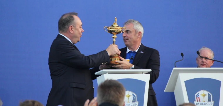 Two men in suits hold the gold Ryder Cup, topped by the small figure of a golfer, between them against a blue backdrop at the closing presentation (2014).