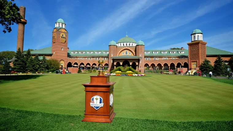 The gold Ryder Cup trophy on a wooden plinth on a striped lawn in front of the brick clubhouse at Medinah Country Club under a blue sky (2012).
