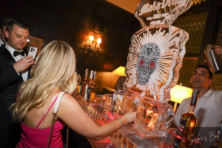 A guest at the tequila ice luge carved with a sugar skull, a bartender raising a shaker beside it, at The Sailors Ball in New York, an April engagement in the season calendar of The Mansa Court
