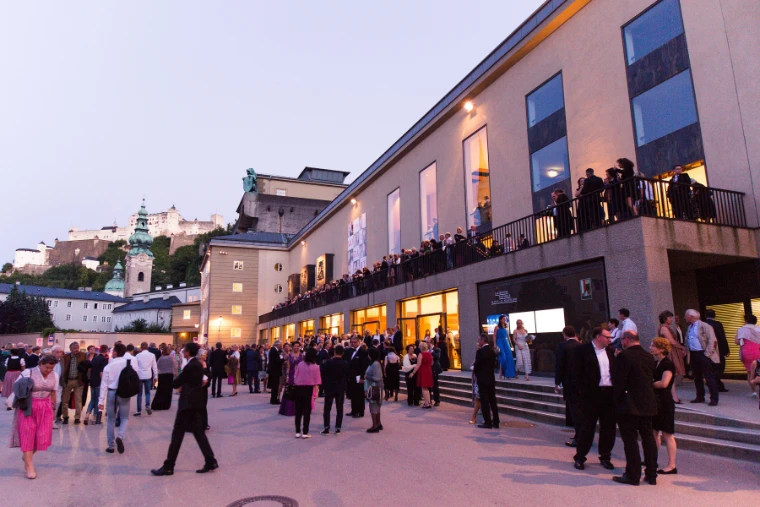 Festival-goers in evening dress gather outside the Grosses Festspielhaus at dusk, the fortress on the hill behind.