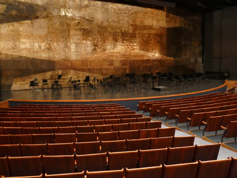 Rows of red seats in an empty auditorium sweep toward a wide stage backed by a gold-leaf wall, with orchestra chairs set out (2018).