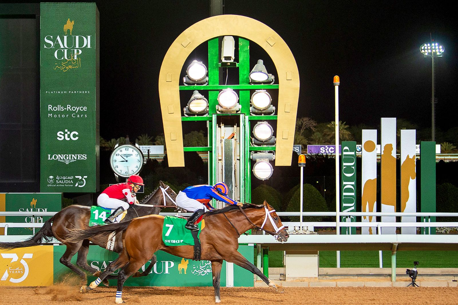 Two horses passing the winning post beneath the golden horseshoe and Longines clock under floodlight at The Saudi Cup