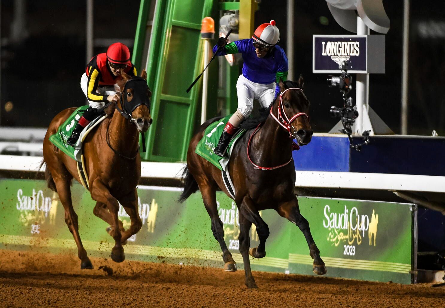 A jockey in blue and green silks raising his whip as he crosses the line ahead of the field at The Saudi Cup