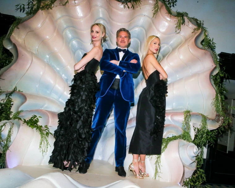 Three guests in a velvet dinner jacket and black gowns posing inside a giant scallop shell hung with greenery at The Save Venice Ball in New York, a late-April engagement in the season calendar of The Mansa Court