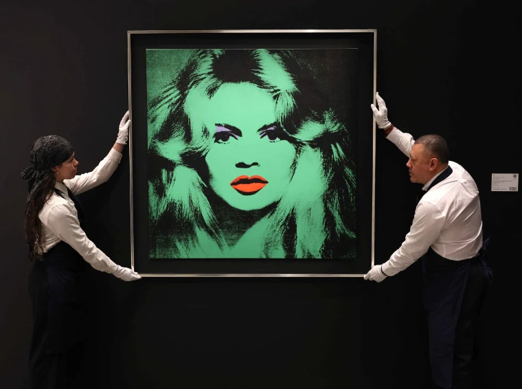 Two art handlers in white gloves present a green Warhol portrait on the viewing wall before the sale. The spring auctions in New York, in the season calendar of The Mansa Court