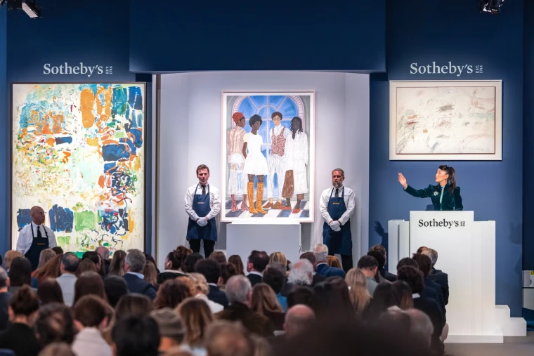 The auctioneer points to a bid from the rostrum at a Sotheby’s evening sale, handlers in aprons flanking the lot. The spring auctions in New York, in the season calendar of The Mansa Court