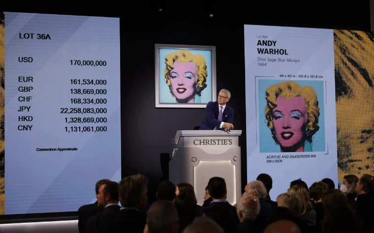The auctioneer waits at the rostrum at Christie’s as a Warhol Marilyn sells, the price shown in seven currencies, in 2022. The spring auctions in New York, in the season calendar of The Mansa Court