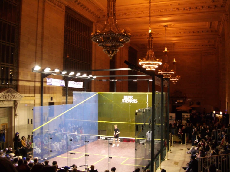 A glass-walled squash court lit under the chandeliers of Vanderbilt Hall, with spectators around it and players on court (2005).