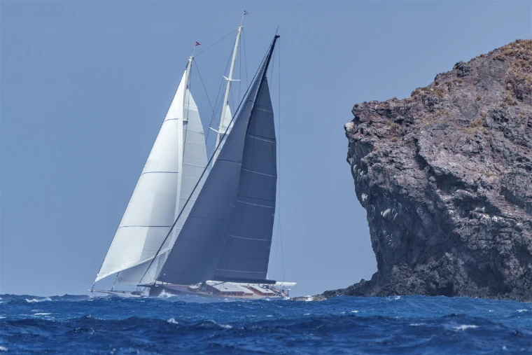 A superyacht under full sail passes a rocky islet off St. Barths (2023). The St. Barths Bucket, off Gustavia, St. Barthélemy, in the season calendar of The Mansa Court