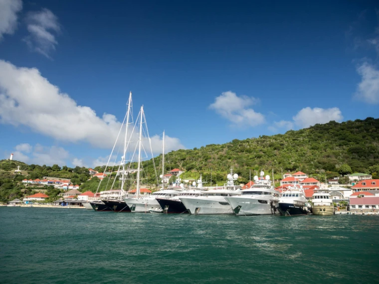 Superyachts moored along the quay of Gustavia harbour under a blue sky, white sailing masts and red-roofed houses on the green hill behind (2013).