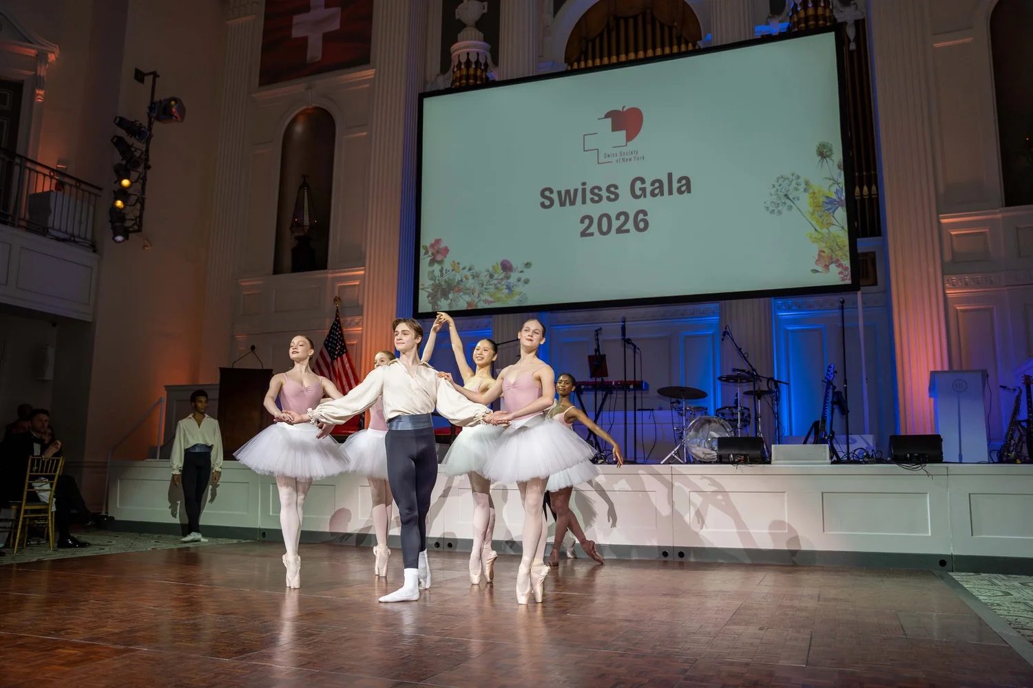 Dancers en pointe performing on stage beneath the Swiss flag at The Swiss Ball, the New York gala The Mansa Court keeps each March