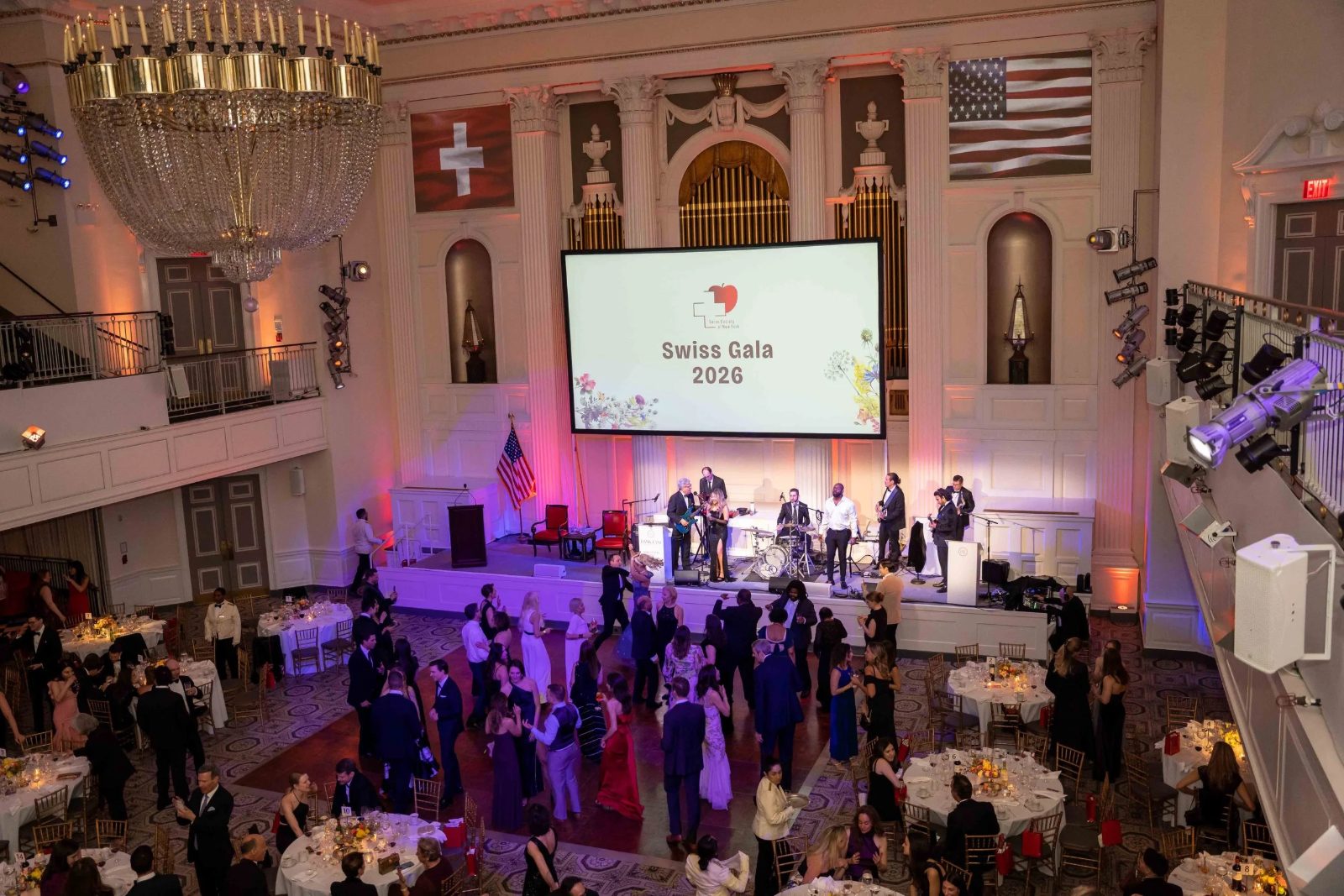 The ballroom seen from the balcony at The Swiss Ball, guests dancing before the band beneath the Swiss and American flags, the March black-tie evening in New York kept in The Mansa Court calendar