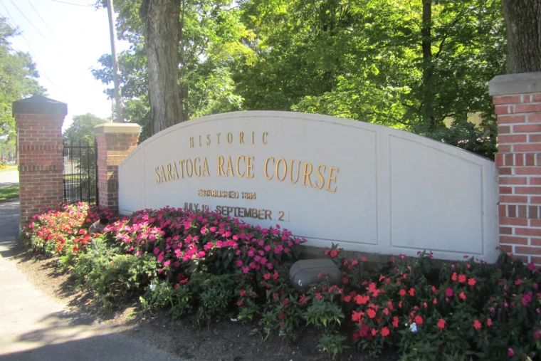 The curved stone sign reading Historic Saratoga Race Course stands between brick pillars behind a bed of pink and red flowers, shaded by trees (2013).