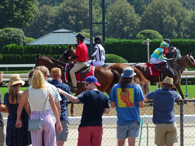 Spectators in summer hats and shirts stand at the rail as jockeys on thoroughbreds, led by outriders, pass in front of green trees (2021).