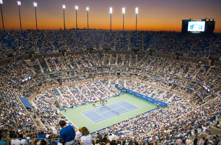 A packed Arthur Ashe Stadium seen from the upper tier at sunset, floodlights glowing over the blue hard court far below (2008).