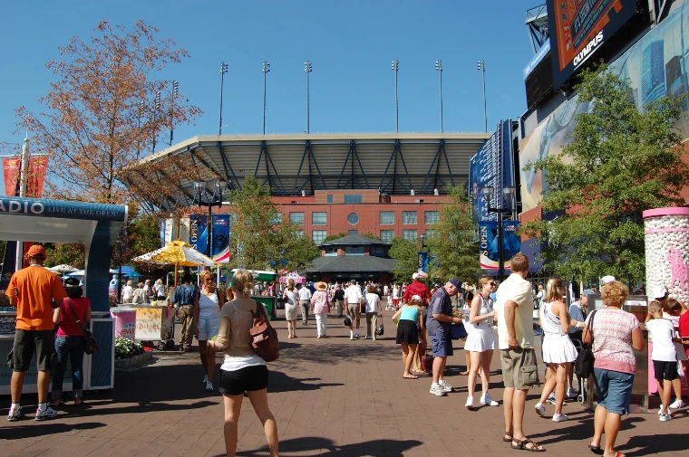 Spectators in summer clothes stroll a sunny plaza on the tournament grounds toward the red-brick facade of Arthur Ashe Stadium (2008).