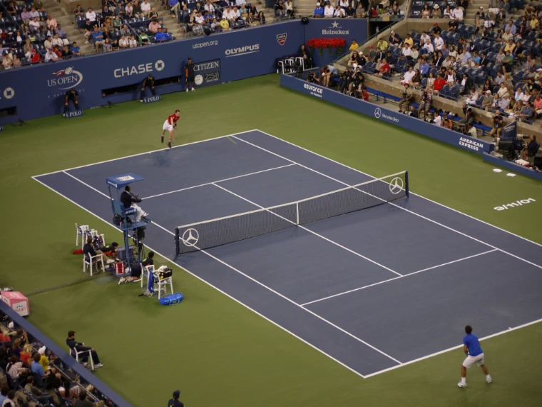 Two players at opposite baselines of a blue hard court during a match under floodlights, with the umpire’s chair and crowded stands alongside (2011).