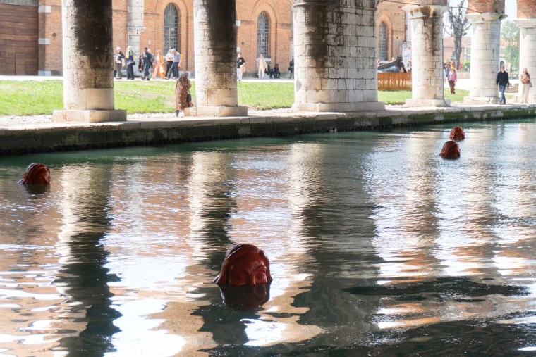 Red sculpted heads by Alice Maher rising from the water of the Arsenale basin beneath its stone colonnade at the Venice Biennale, an engagement in the season calendar of The Mansa Court