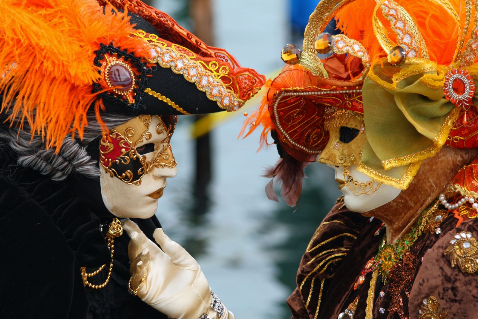 Two carnival masks in gilt and orange plumes turned face to face above a canal at The Venice Carnival