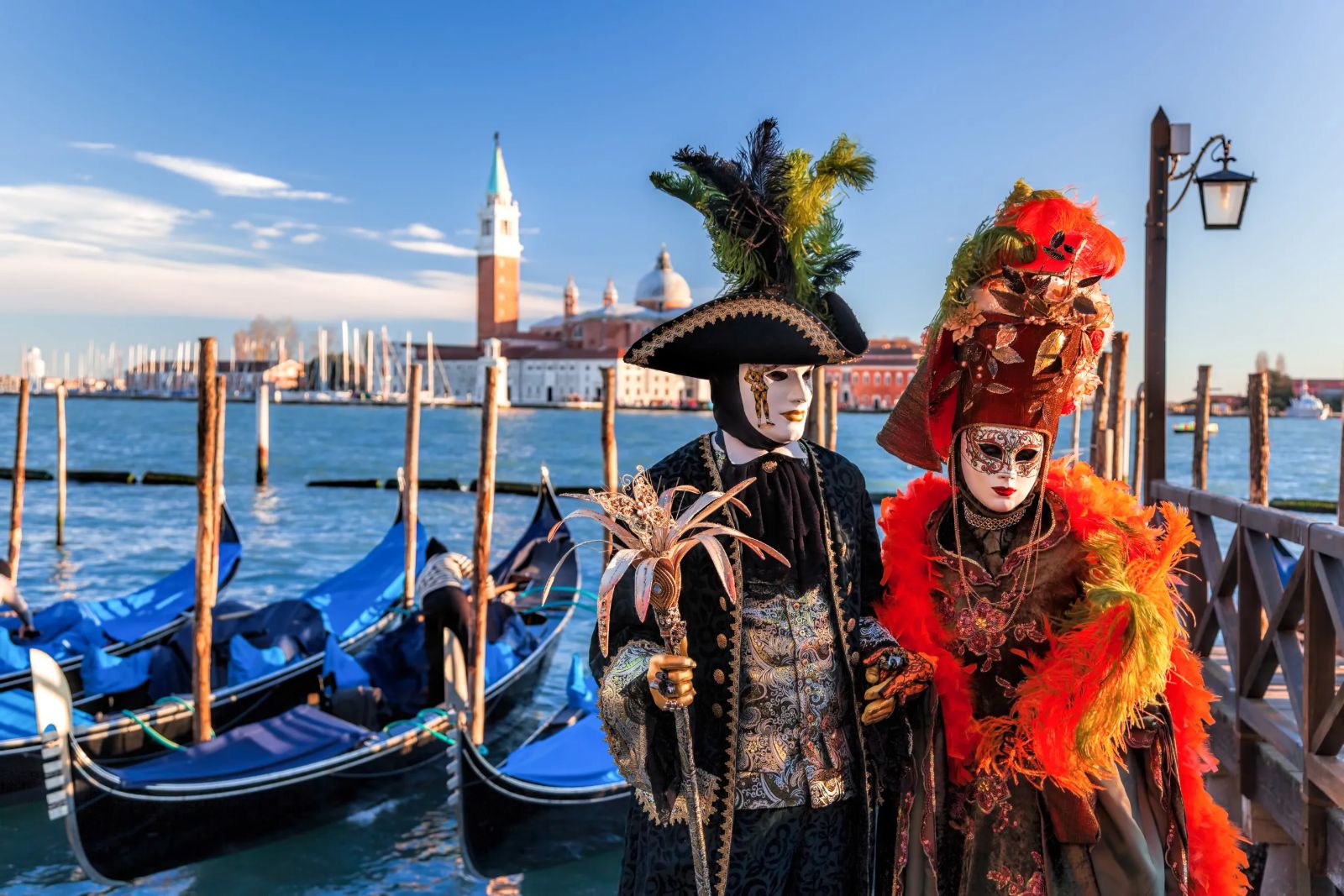 A masked pair in tricorne and plumed headdress on the Riva above the moored gondolas with San Giorgio Maggiore beyond at The Venice Carnival