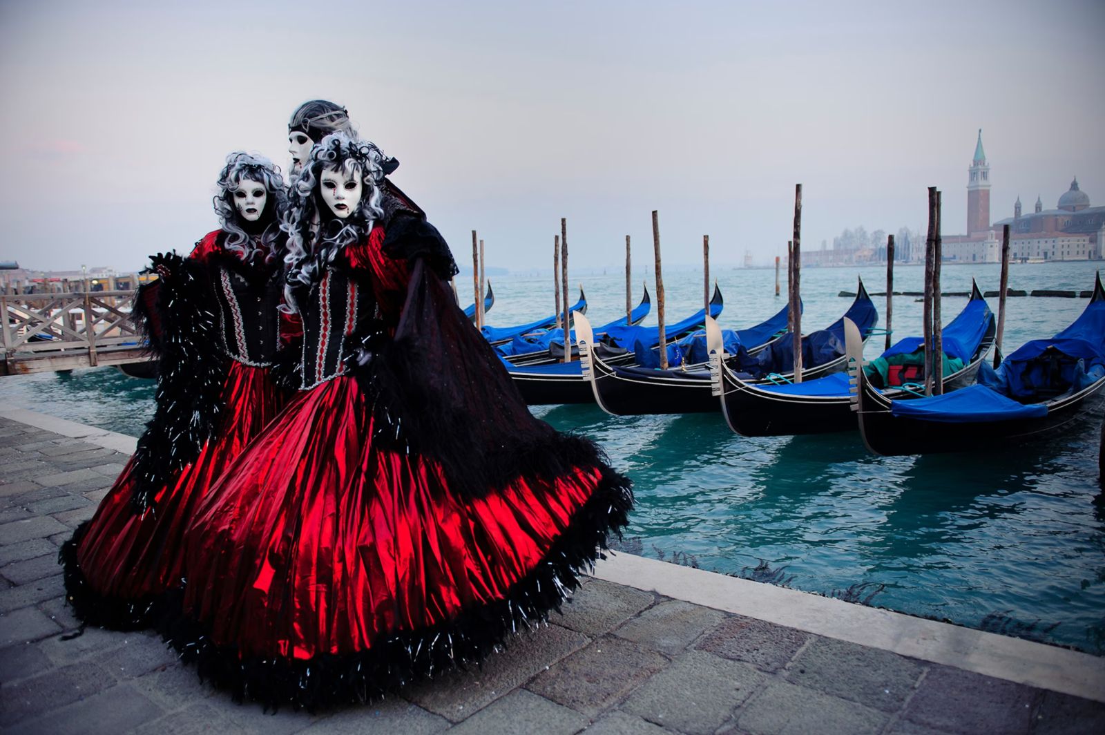 Three costumed figures in red and black silk with white masks and powdered wigs beside the moored gondolas at The Venice Carnival