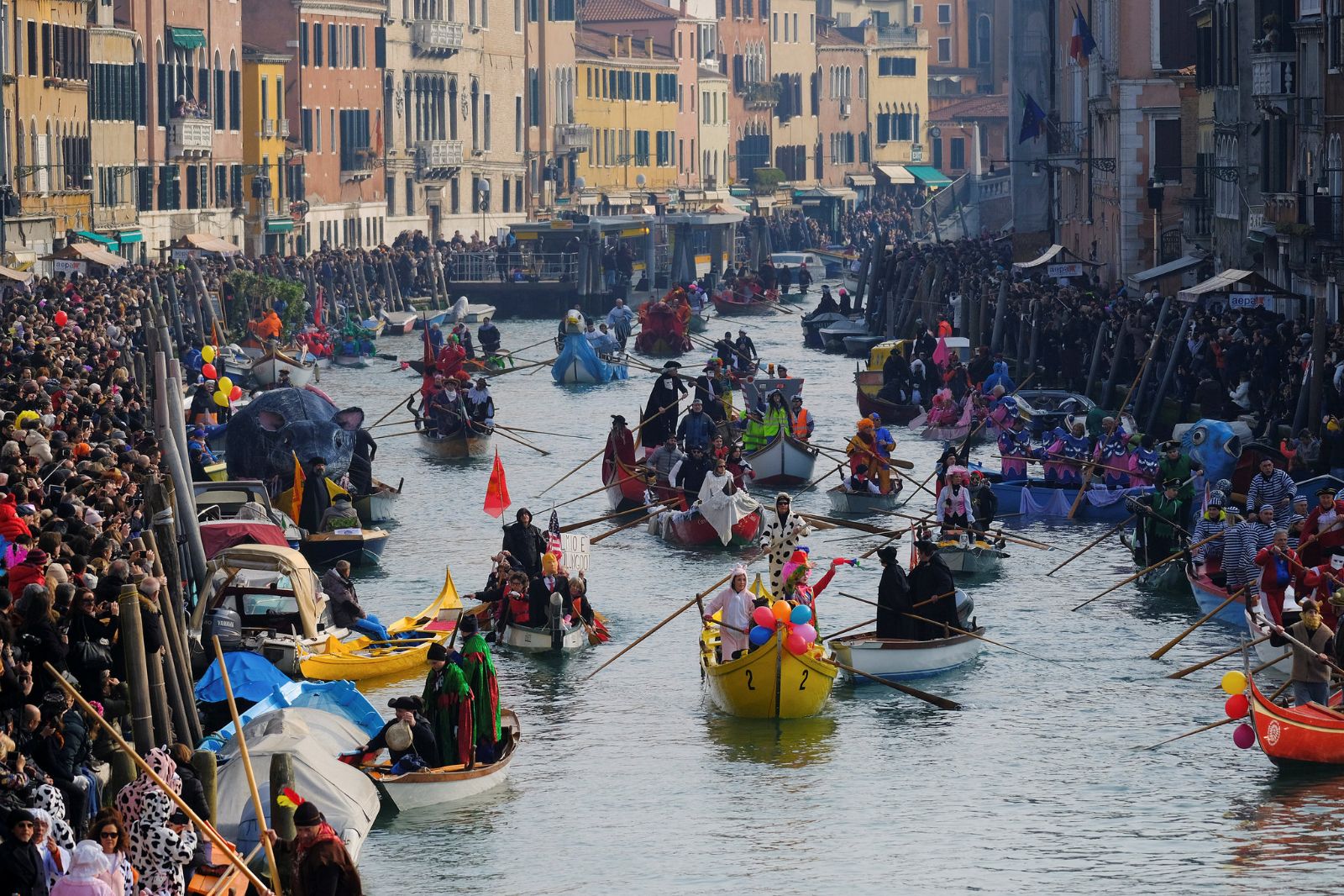 A flotilla of costumed rowers and decorated boats filling the canal between crowded banks at the water parade of The Venice Carnival