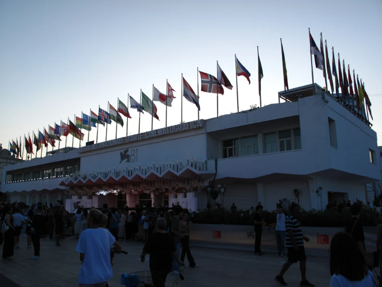 The white Palazzo del Cinema with a long row of national flags on its roof at dusk, with festivalgoers gathered on the forecourt (2024).