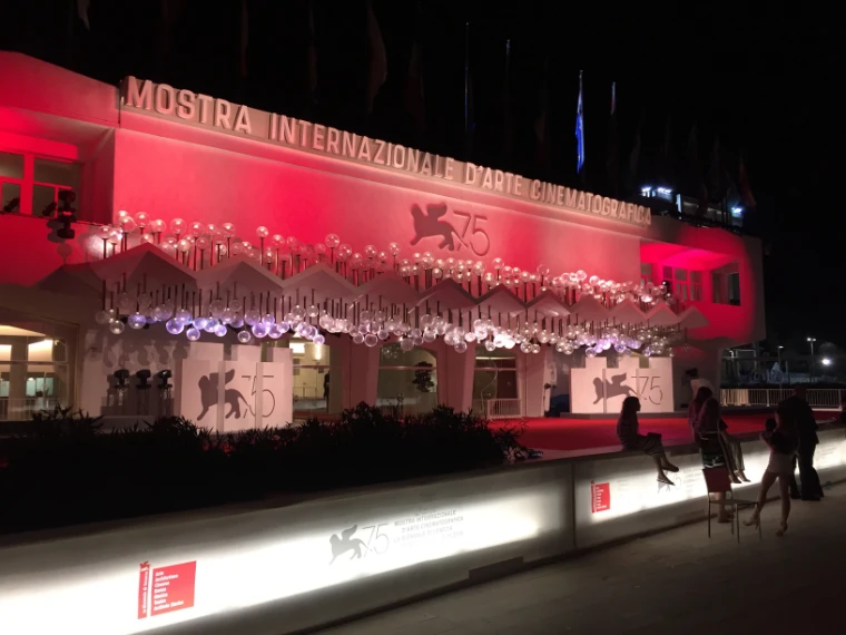 The facade of the Palazzo del Cinema glows red at night above rows of glass globe lights and the 75th Mostra lion emblem (2018).