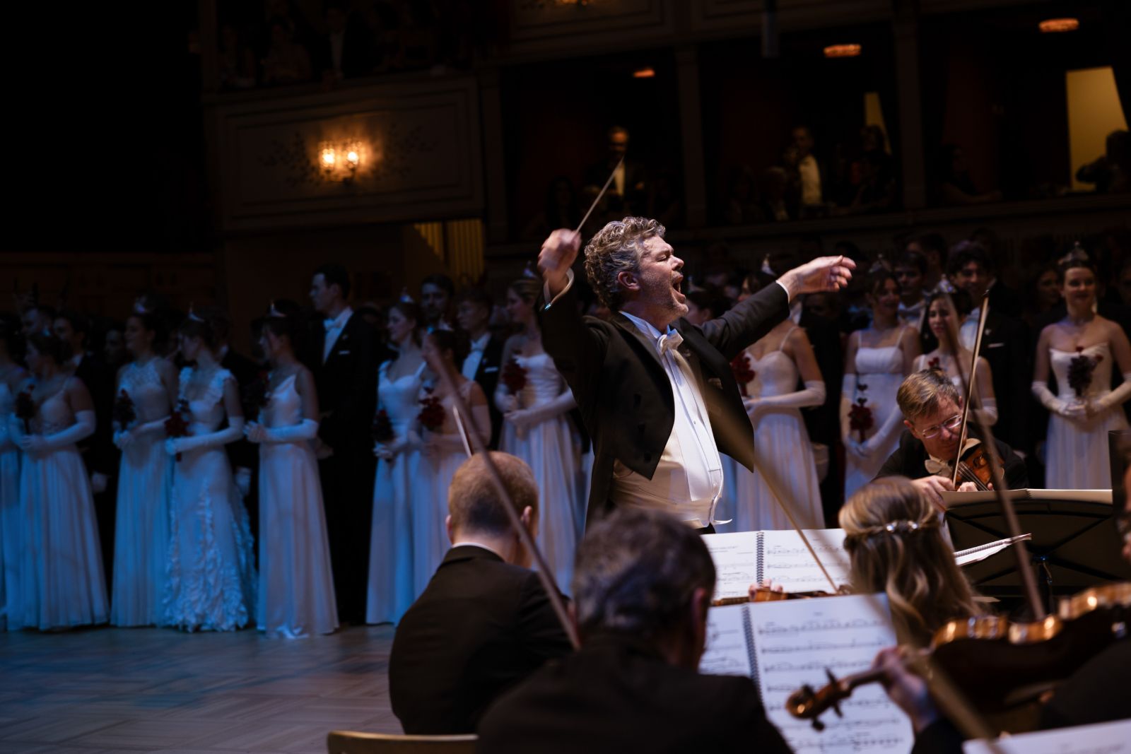 The conductor at full stretch on the podium with debutantes ranked in white behind him at The Vienna Opera Ball