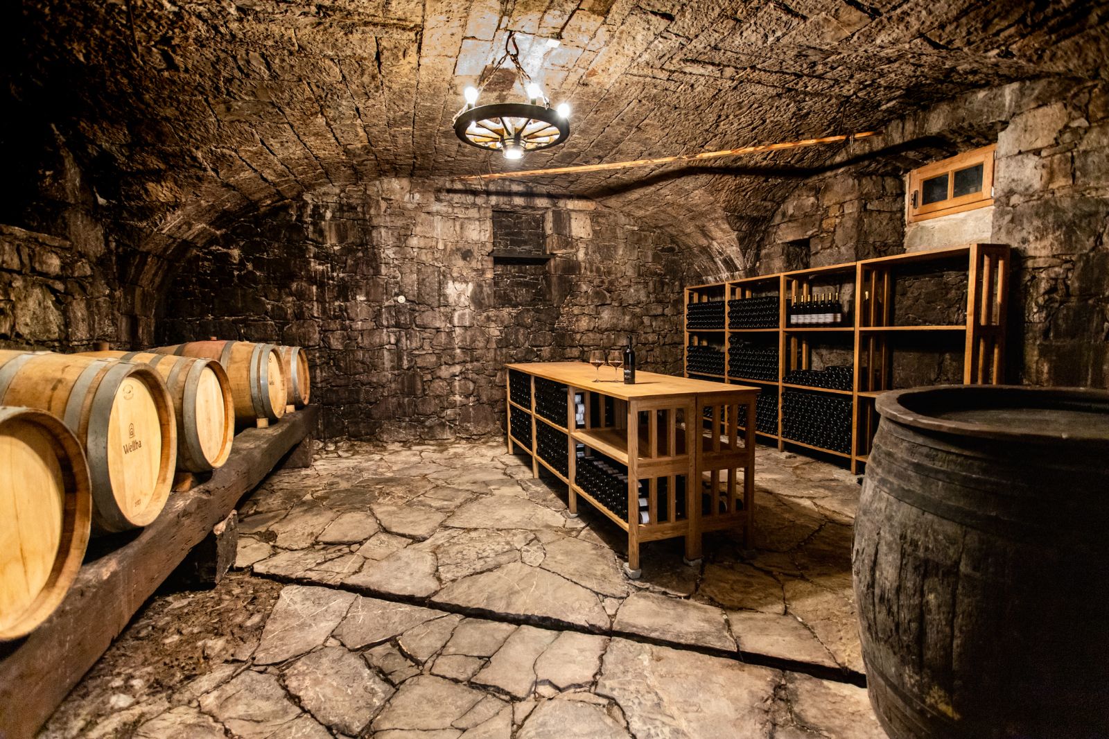 A vaulted stone cellar with oak barrels along one wall, bottle racks against the back, and a wooden tasting table under an iron chandelier