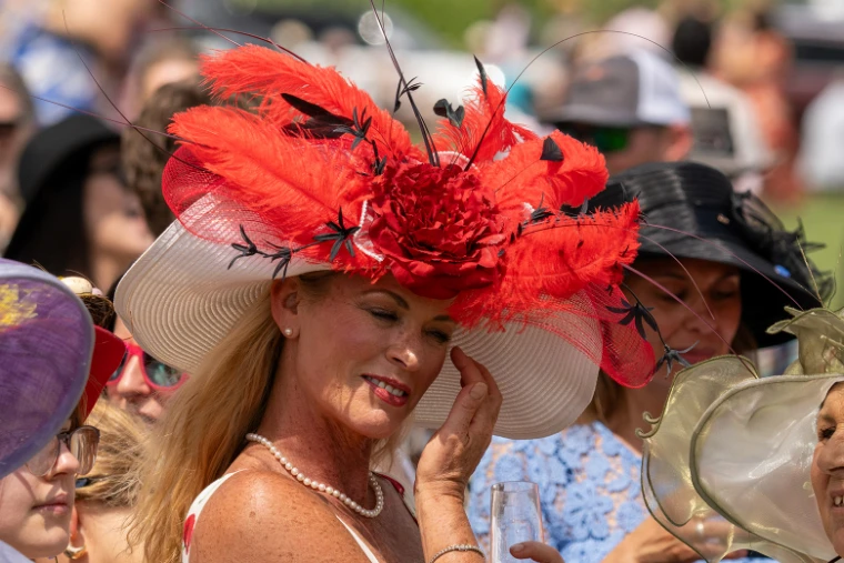 A racegoer in a wide white hat crowned with red plumes and a silk peony, pearls at her throat, in 2025. The Virginia Gold Cup at Great Meadow, The Plains, Virginia, in the season calendar of The Mansa Court