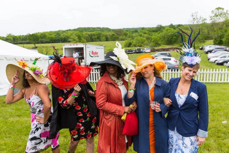 Five racegoers in flowered and feathered hats pose arm in arm on the grass above the course. The Virginia Gold Cup at Great Meadow, The Plains, Virginia, in the season calendar of The Mansa Court