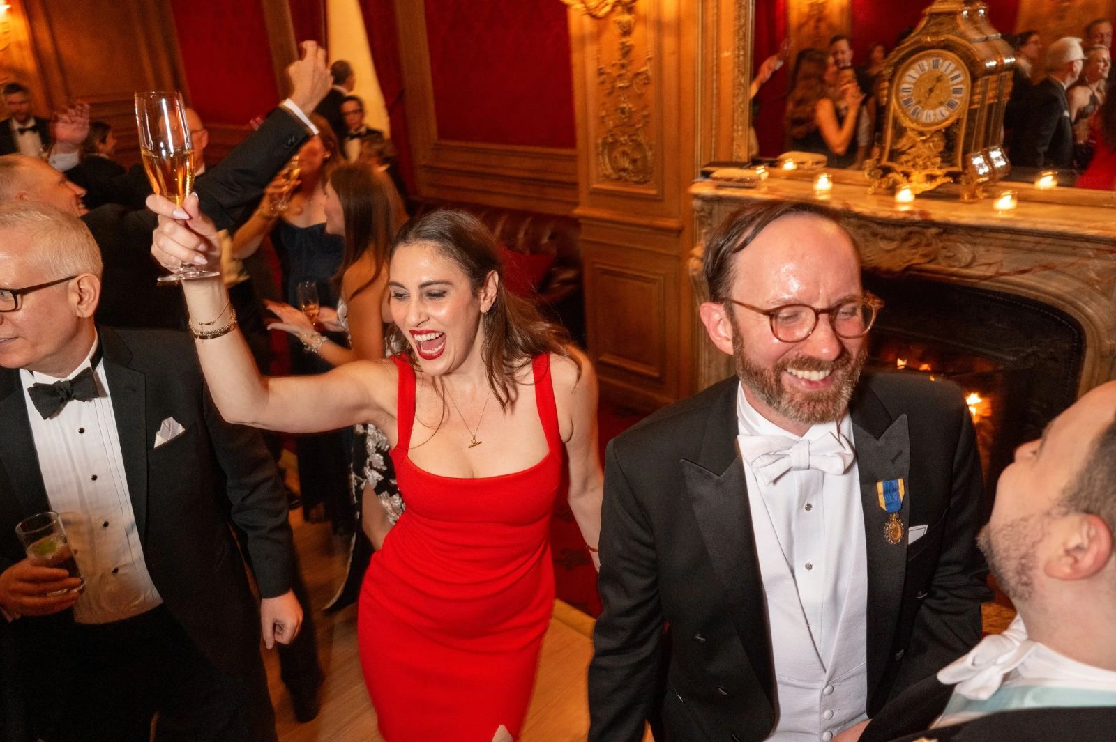 Guests dancing with raised champagne before the fire in a panelled crimson room at The Washington Birthday Ball