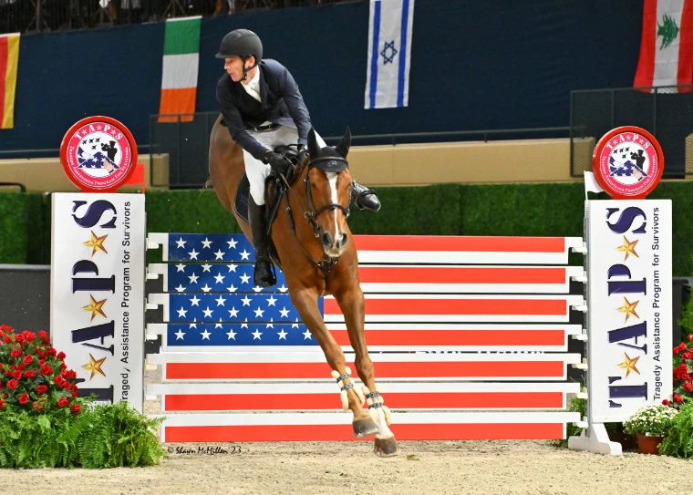 A chestnut horse and rider jumping a fence painted as the American flag, flanked by TAPS standards and red flowers (2023).