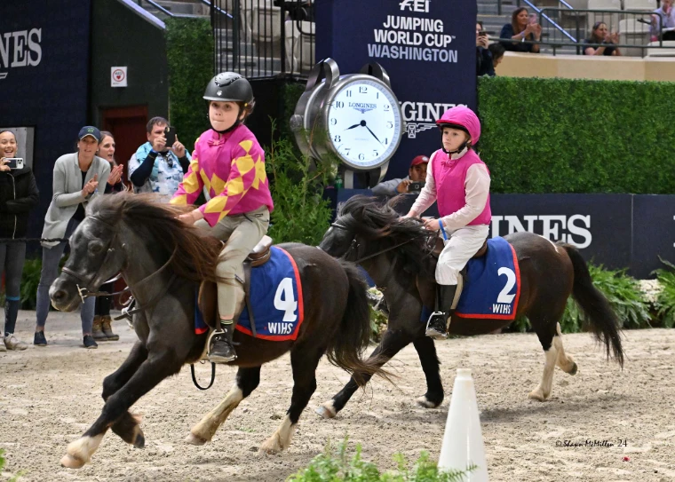 Two young jockeys in bright silks racing Shetland ponies across the arena sand past a giant clock (2024).
