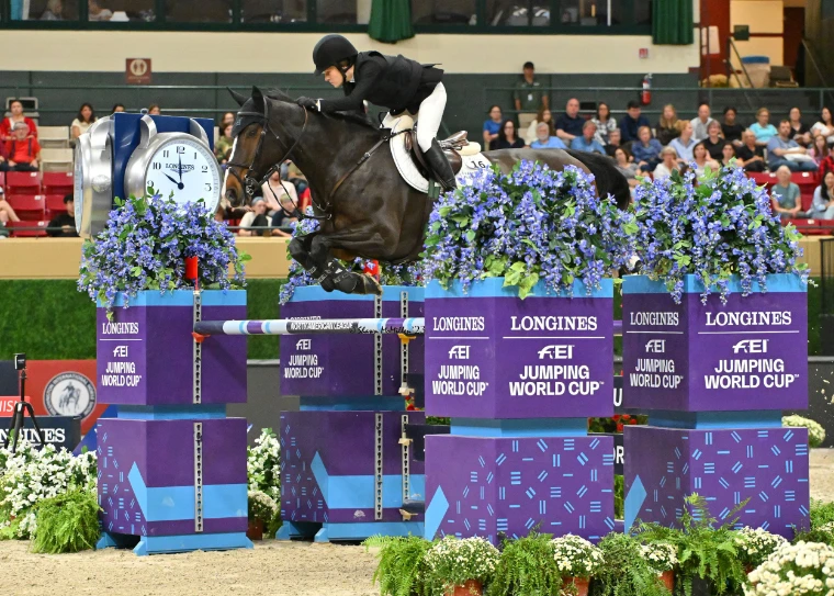 A rider on a dark horse clearing a purple Longines FEI Jumping World Cup oxer dressed with blue flowers, the indoor stands behind (2023).