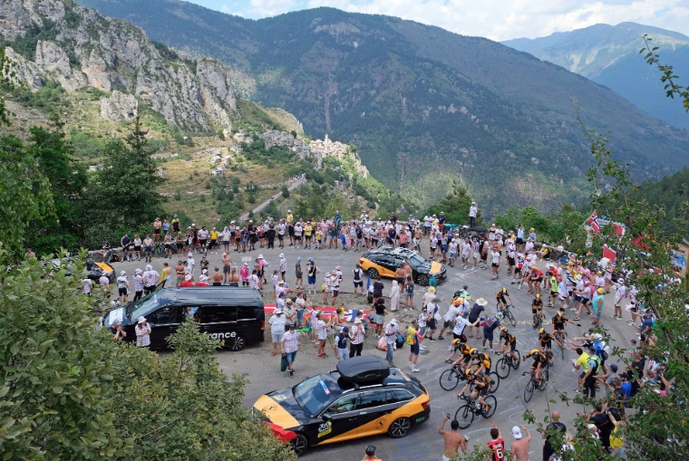 A club group in matching kit climbs through a hairpin on the Col de la Couillole ahead of the race, the crowd packed on the bend (2024).