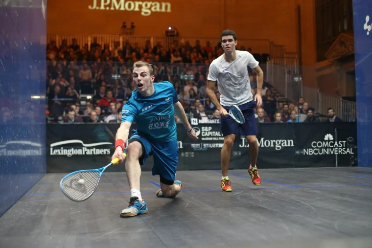A player lunges for a low ball as his opponent watches. Tournament of Champions, Grand Central Terminal, New York (2016), in the season calendar of The Mansa Court