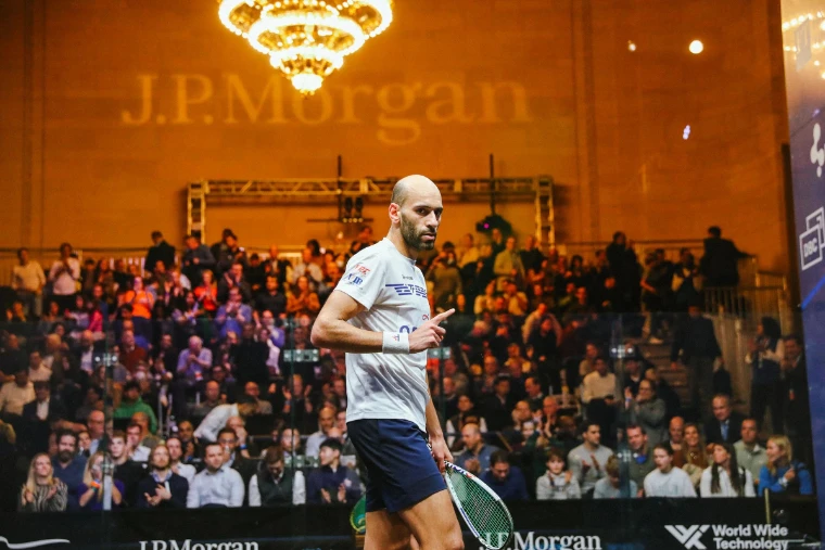 A player in white turns after a point before a packed gallery. Tournament of Champions, Grand Central Terminal, New York, in the season calendar of The Mansa Court