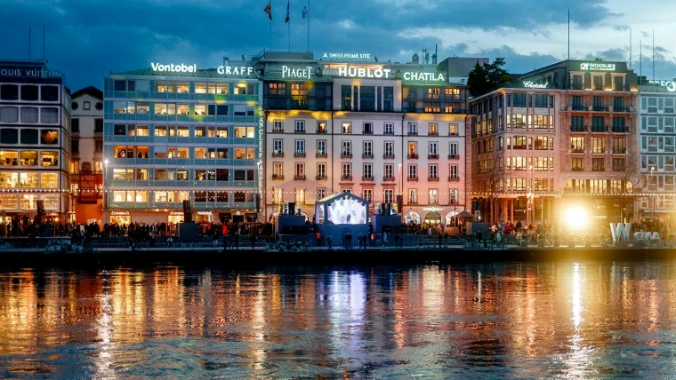The Geneva lakefront at dusk, its facades lit with the names of the watch houses and a stage set on the quay, during the week of Watches and Wonders, a mid-April engagement in the season calendar of The Mansa Court